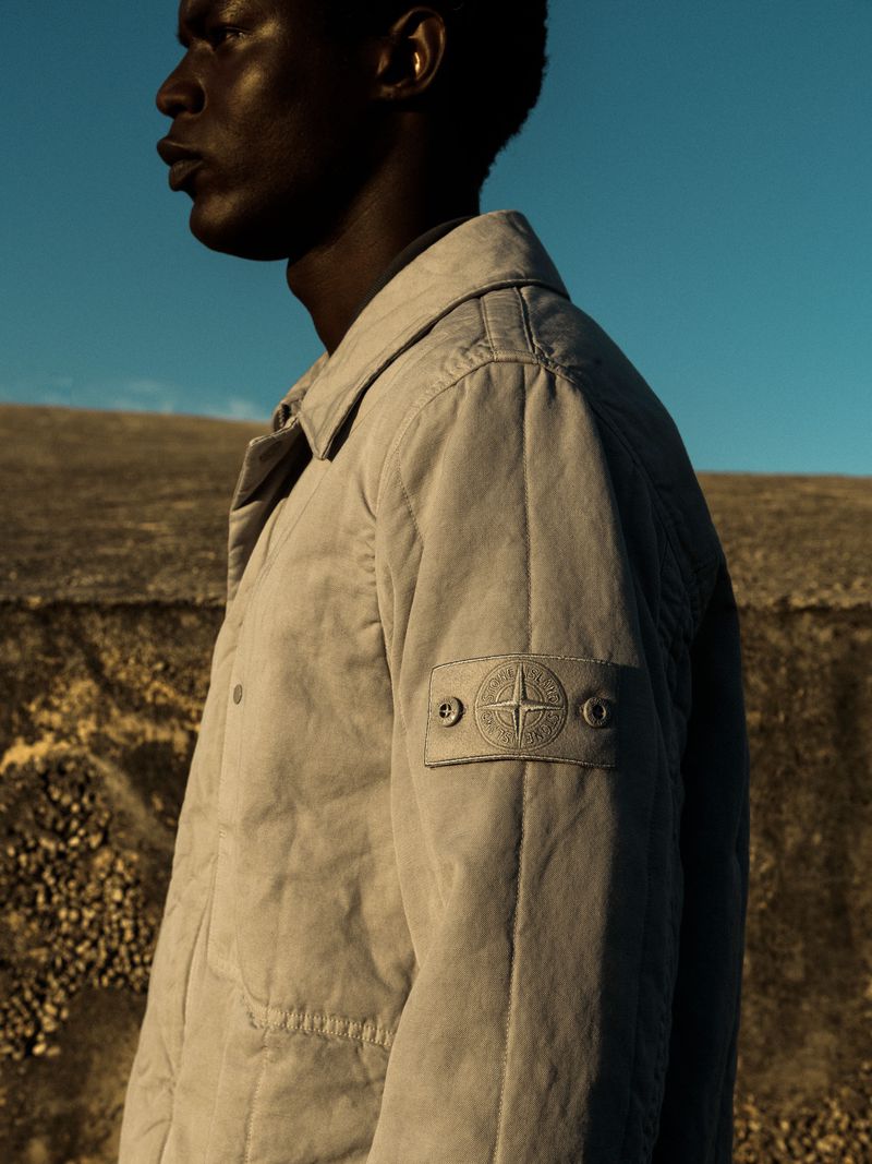 Profile view of a black man wearing a light beige jacket with a sleeve badge against an outdoor landscape and blue sky