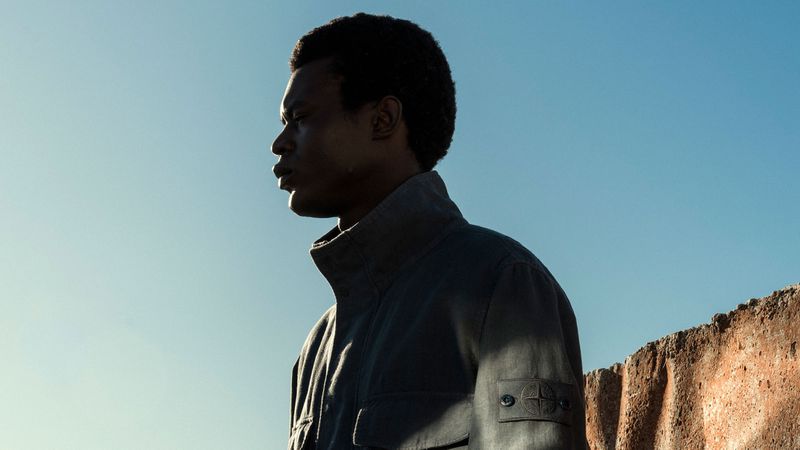 Profile portrait of a black man wearing a dark jacket with a compass patch against a clear blue sky.