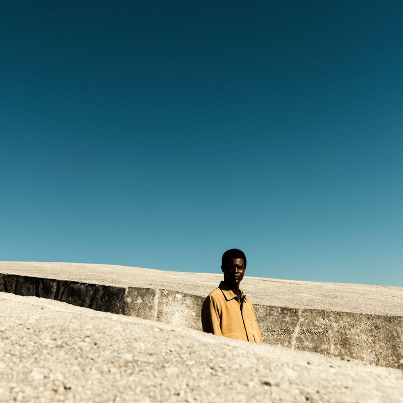 African American man in beige jacket poses against a pale concrete structure under a bright blue sky on a sunny day.