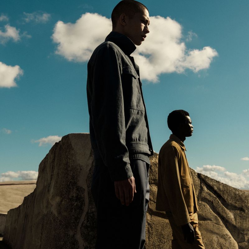 Two men standing against a rocky outdoor backdrop under a blue sky with scattered white clouds for Stone Island.