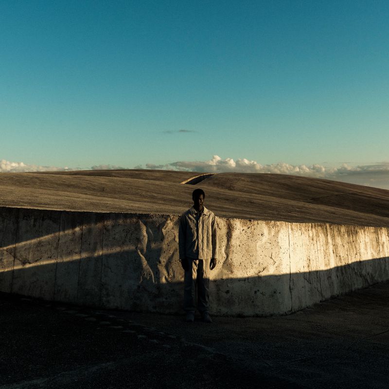 A man standing before a textured concrete wall under a blue sky, photographed by Christopher Anderson for Stone Island.