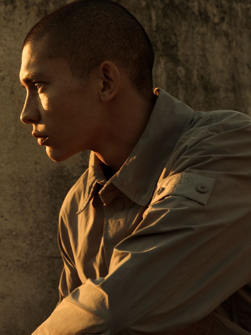 Profile portrait of a young man with a buzz cut wearing a buttoned jacket in warm golden light against a textured concrete wall