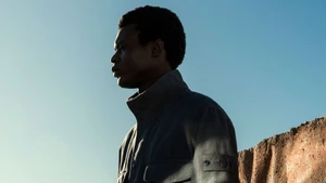 Profile portrait of a black man wearing a dark jacket with a compass patch against a clear blue sky.