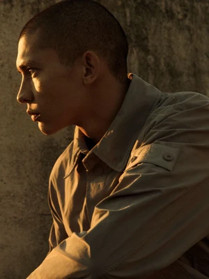 Profile portrait of a young man with a buzz cut wearing a buttoned jacket in warm golden light against a textured concrete wall