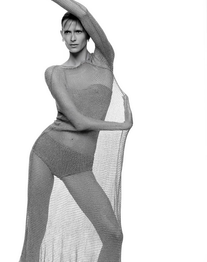 Monochrome portrait of a slender adult female fashion model in a draped metallic mesh gown, posing in a white photo studio.