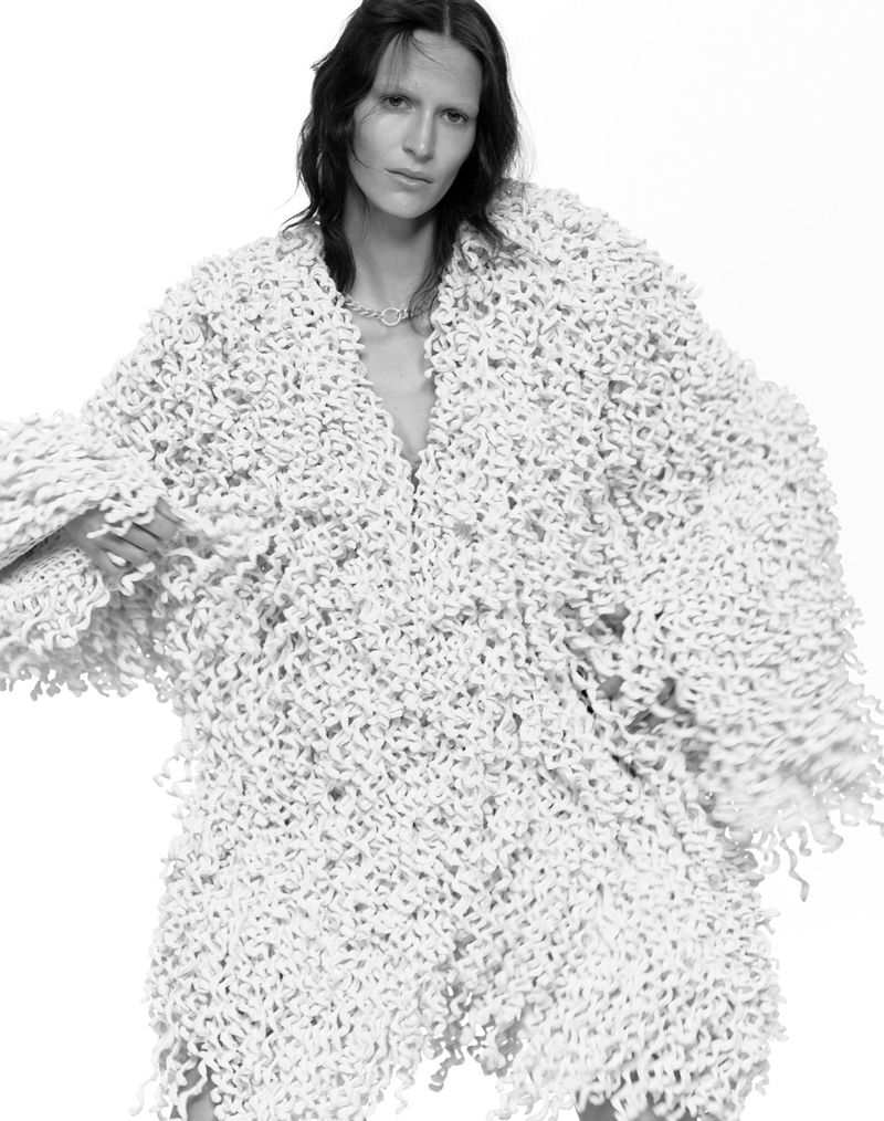 Monochrome portrait of a slender woman wearing a unique dress with spiral textures, set against a stark white backdrop in a studio.