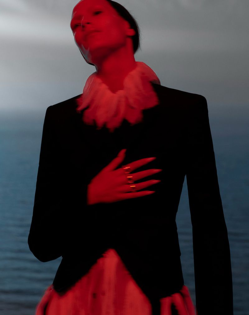 Fashion portrait of a woman in a black blazer and white ruffled collar against a blurred background of the sky and sea.