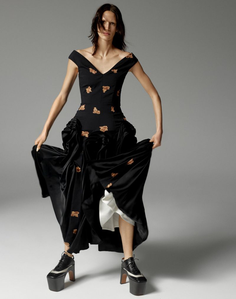 Fashion model poses in a black floral dress with platform shoes against a gray studio backdrop, showcasing editorial style and design.