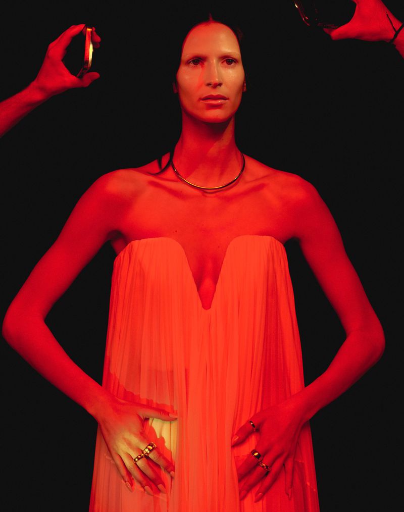 Adult female in a strapless dress poses in studio setting with red lighting and hands holding mirrors to reflect light.