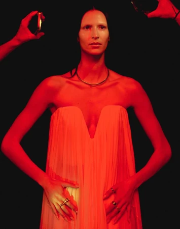 Adult female in a strapless dress poses in studio setting with red lighting and hands holding mirrors to reflect light.