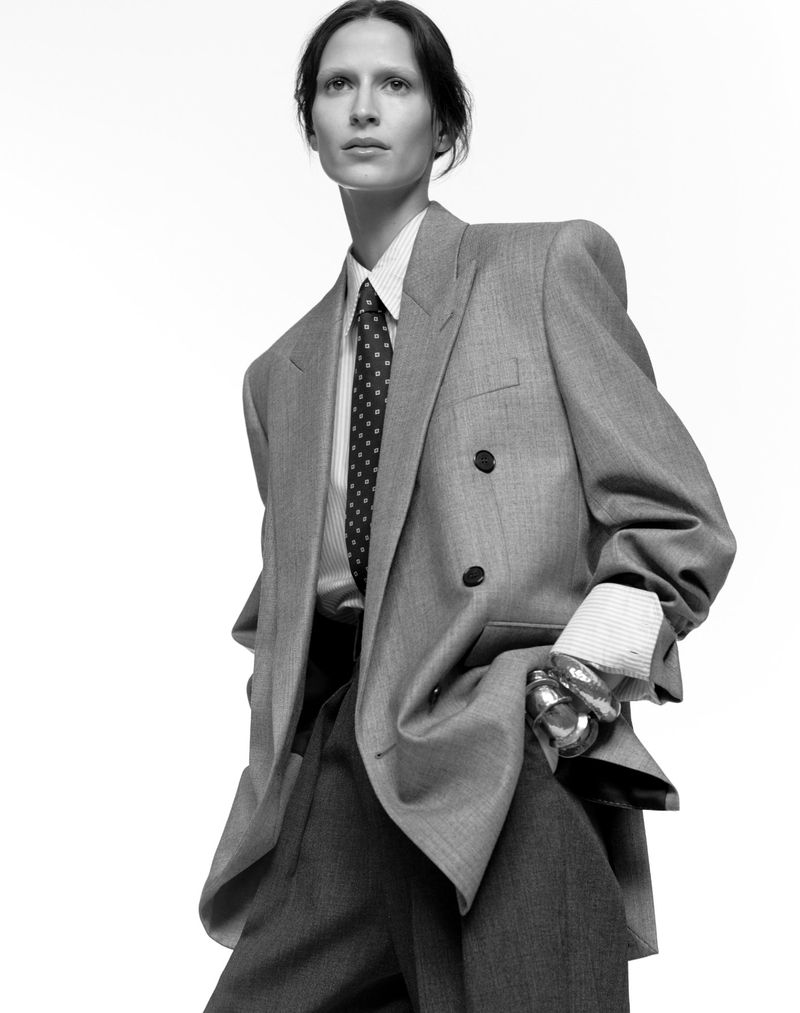Model Jeanne Cadieu wearing an oversized grey double-breasted blazer, shirt, and dotted necktie against a plain white background.