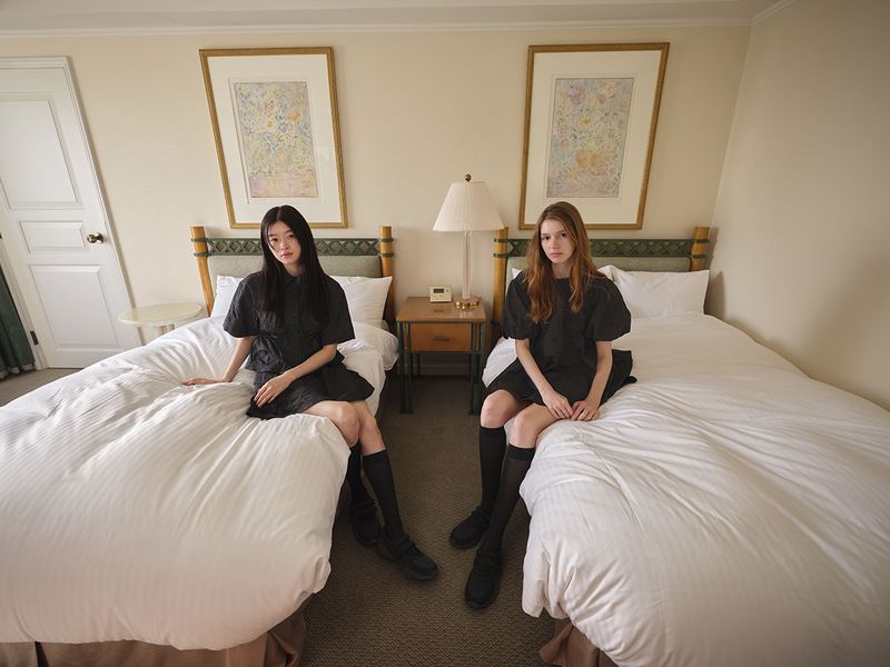 Two female models seated on twin beds in a hotel room wearing matching dark dresses and black knee-high socks.