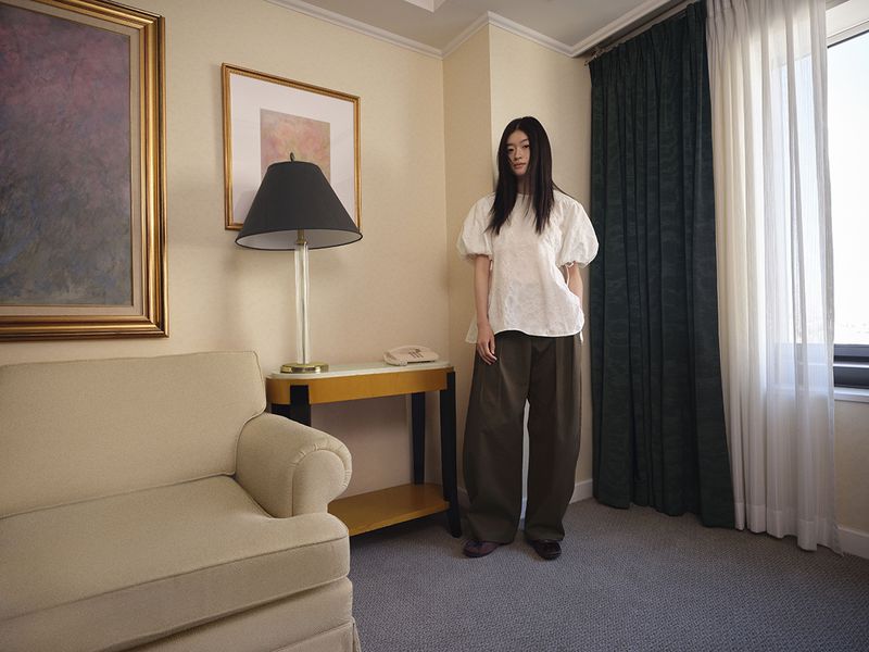 Woman with long dark hair wearing white puff-sleeve top and dark trousers in a hotel room