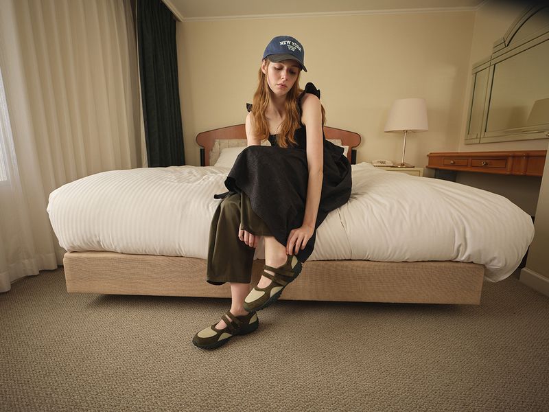 Model wearing a black dress and baseball cap seated on a hotel bed in the GU x Rokh Spring/Summer 2026 collection.