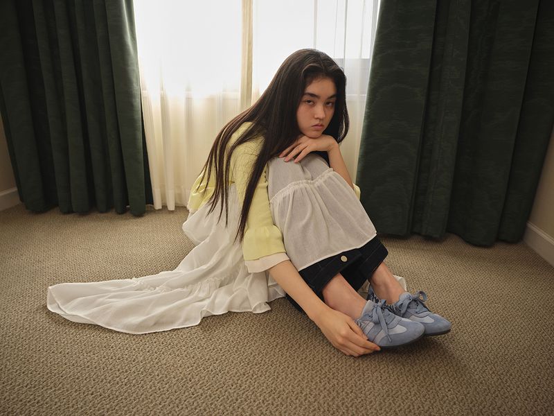 Model seated on a carpeted floor wearing a yellow cardigan, layered white skirt, and blue sneakers