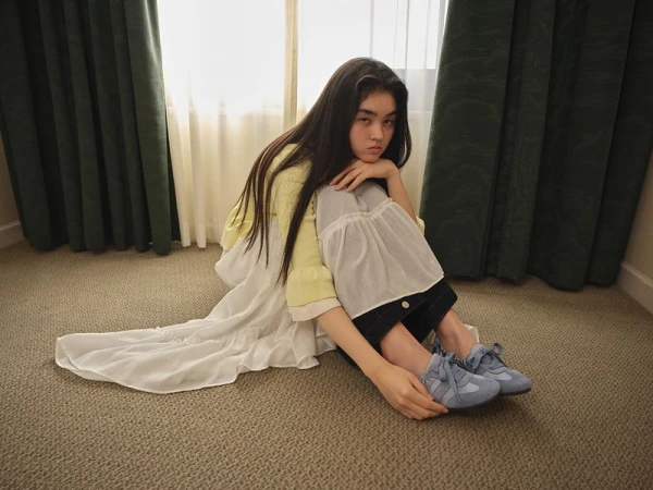 Model seated on a carpeted floor wearing a yellow cardigan, layered white skirt, and blue sneakers
