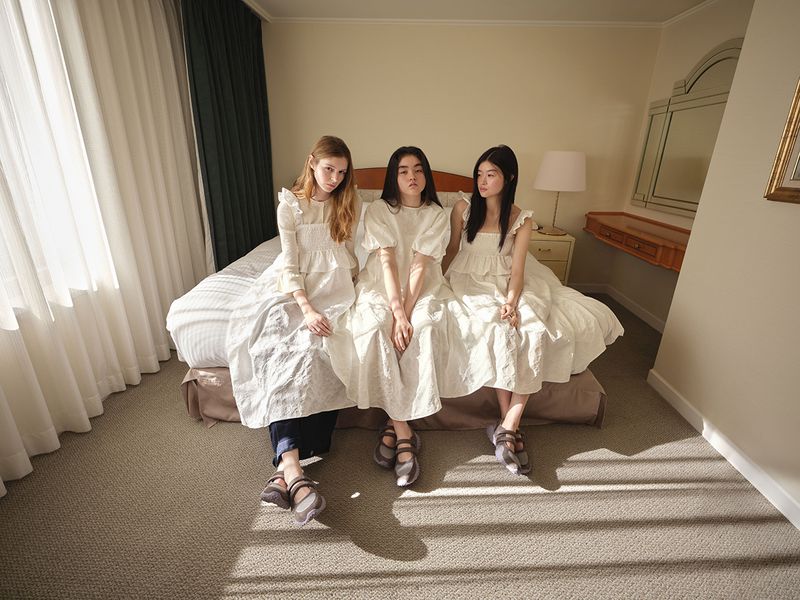 Three models wearing white dresses seated on a hotel bed for the GU x Rokh Spring/Summer 2026 collection.