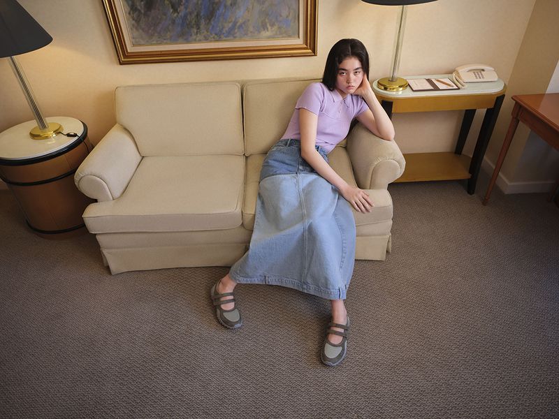 Model wearing a lavender polo shirt and long denim skirt seated on a beige sofa in a hotel room
