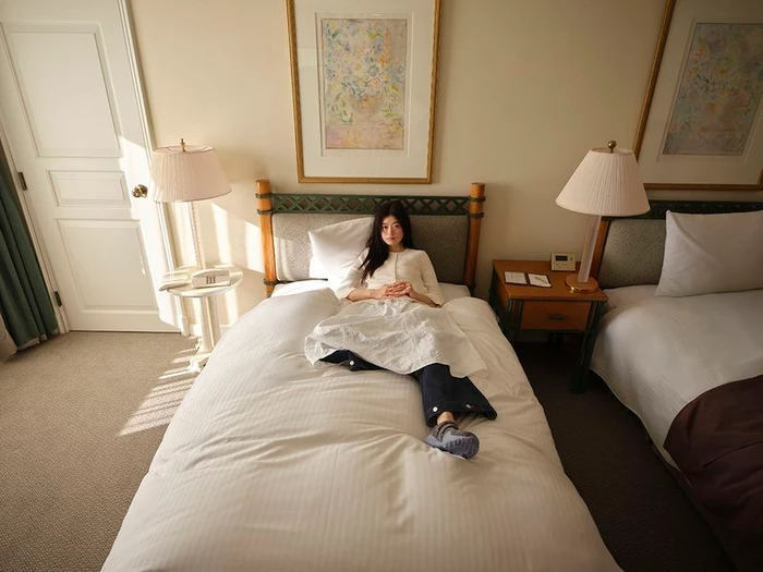 Model lying on a hotel bed in a white top and dark trousers for the GU x Rokh Spring/Summer 2026 collection.