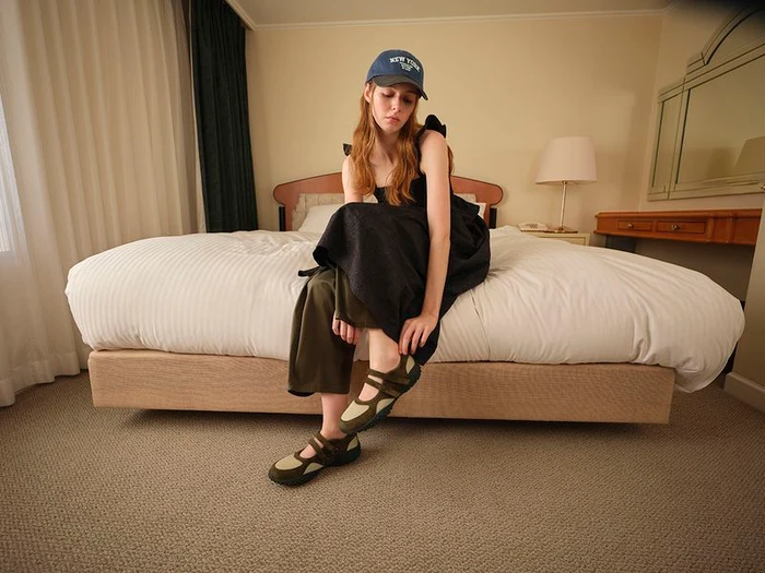 Model wearing a black dress and baseball cap seated on a hotel bed in the GU x Rokh Spring/Summer 2026 collection.