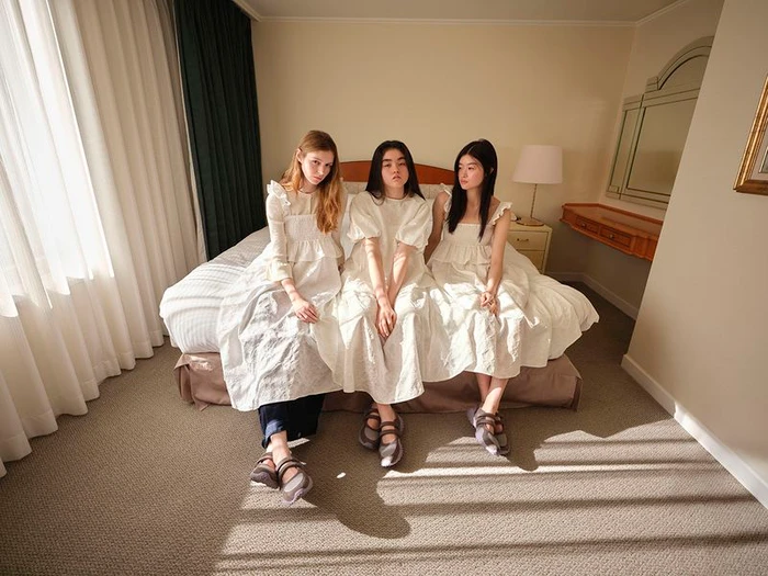 Three models wearing white dresses seated on a hotel bed for the GU x Rokh Spring/Summer 2026 collection.