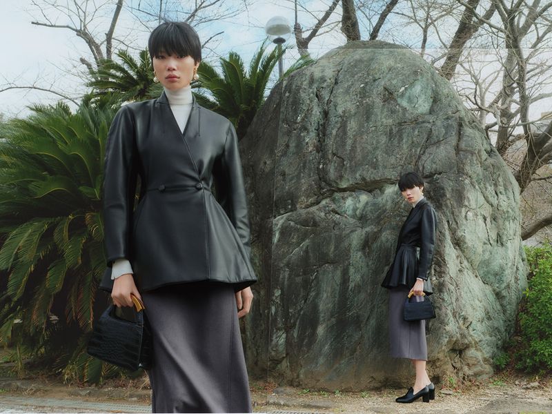 Model in black leather jacket and grey skirt holding a black handbag standing outdoors by a large rock.