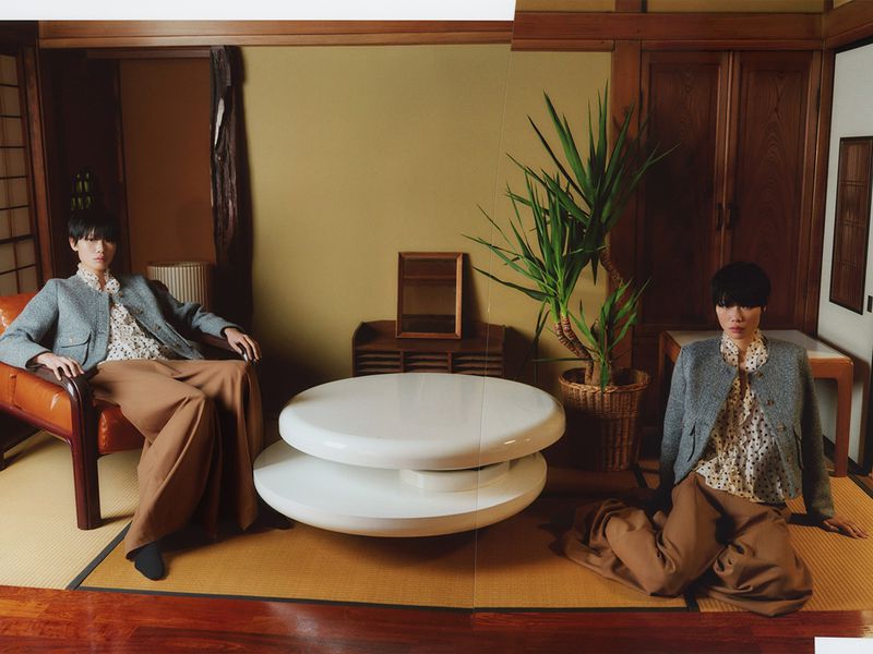 Two models in matching grey jackets and patterned blouses seated in a traditional Japanese room with tatami mats and wooden paneling.