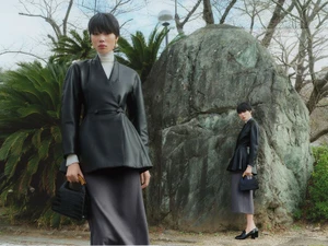 Model in black leather jacket and grey skirt holding a black handbag standing outdoors by a large rock.
