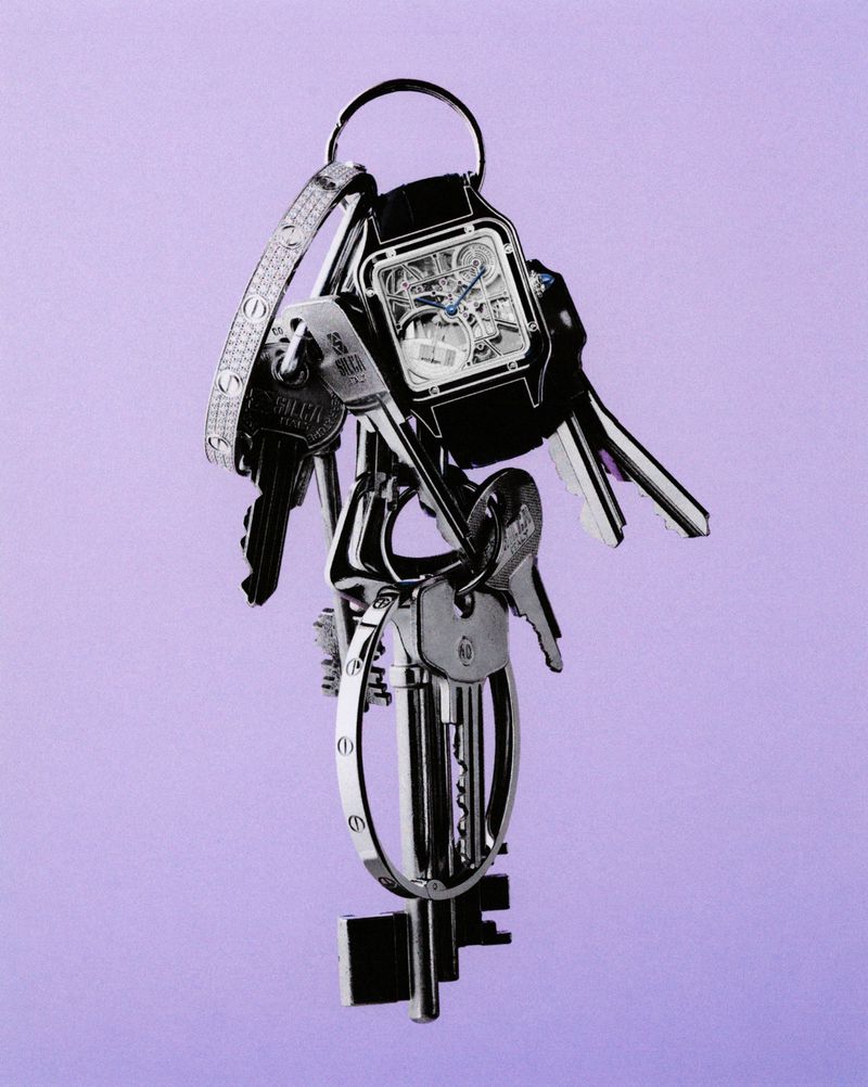 Elevated still life of a black and silver key ring with an open-faced watch, keys, and bangle, on a soft purple backdrop.