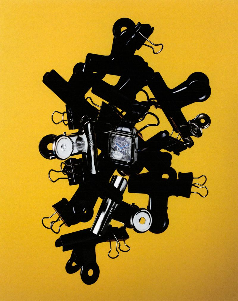 Black binder clips and a wrist watch form a surreal, abstract composition on a solid yellow background with studio lighting.