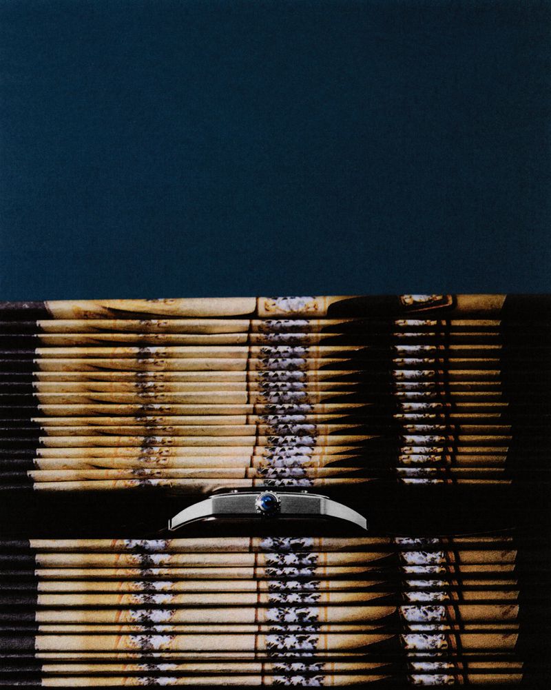 Elevated studio shot: silver wristwatch with a blue gemstone face rests atop stacked brown bamboo window blinds against a deep blue background.