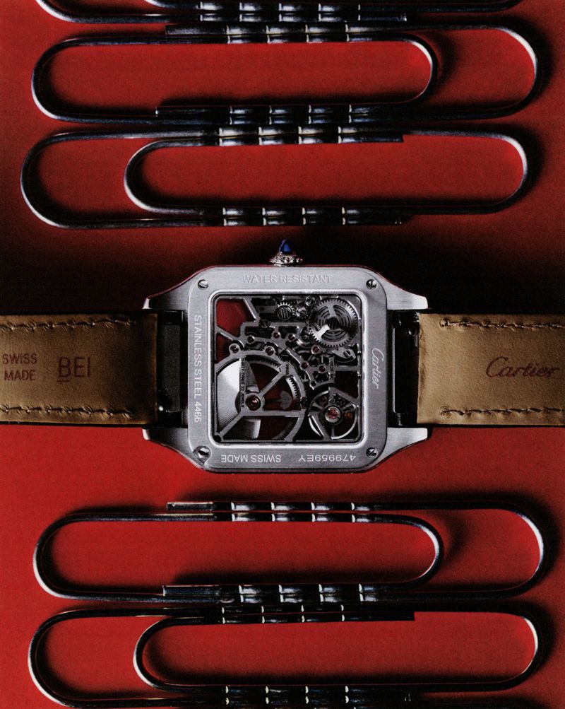 Back of a Cartier watch with visible gears, paired with silver paper clips against a red backdrop, creating a modern, industrial-chic aesthetic.