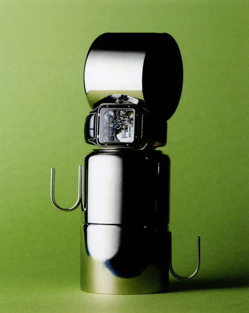 Wristwatch atop a stack of shiny metal cylinders and U-shaped bars against a plain green background.