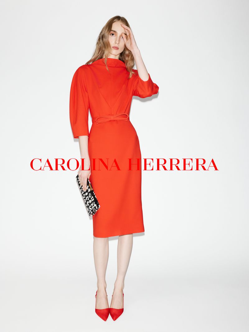 A model in a vibrant red dress and red heels holding a patterned clutch against a plain white studio background.