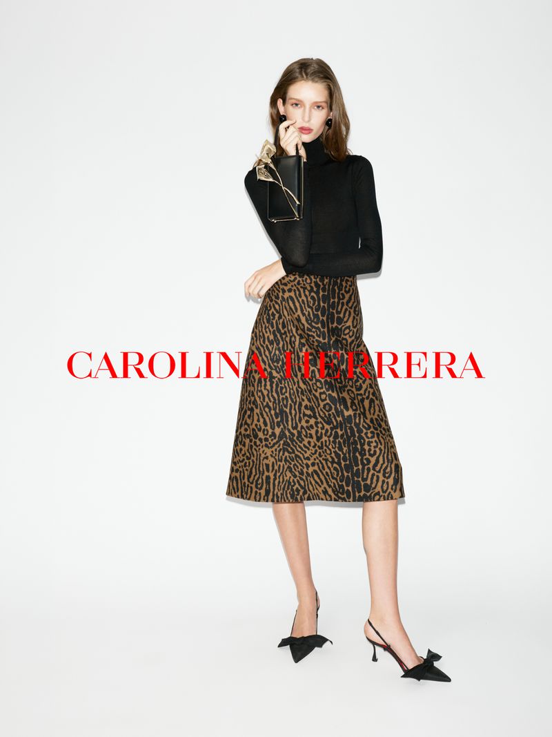 Full-shot of a woman in a black turtleneck and leopard-print midi skirt holding a clutch, posing against a white backdrop.
