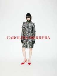 Model wearing a black and white patterned coat, dark sunglasses, and red shoes against a plain white background with red brand text.