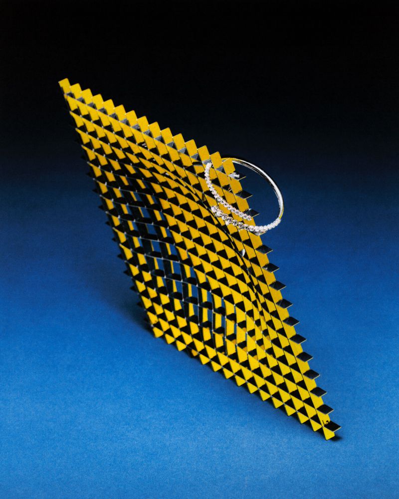 Geometric arrangement with a diamond bracelet. Features a diamond-studded jewelry piece on a textured, yellow and black surface against a gradient blue background.