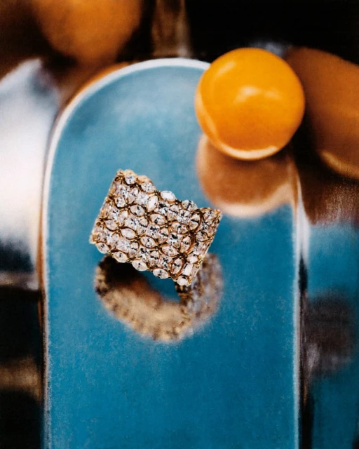 Close-up of a gold and white gemstone encrusted bracelet with an orange sphere, reflected in a shiny teal surface in indoor lighting.