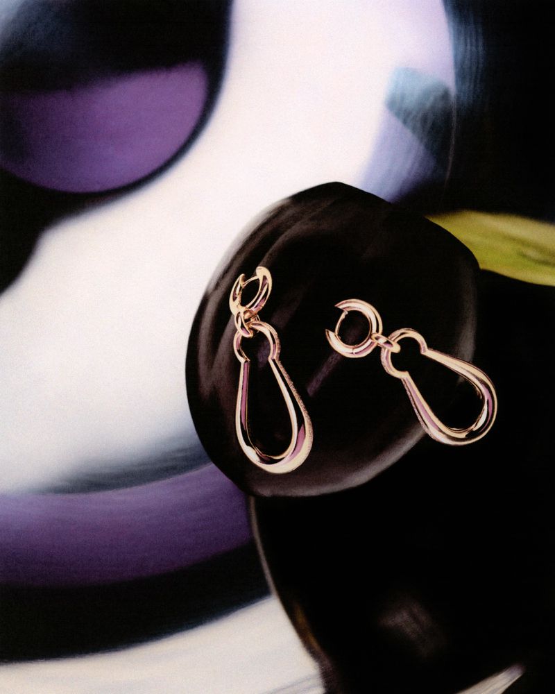 Close-up of gold drop earrings draped on a dark, lustrous surface against a blurred, abstract background of light and dark hues.