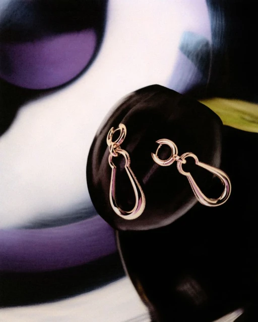 Close-up of gold drop earrings draped on a dark, lustrous surface against a blurred, abstract background of light and dark hues.