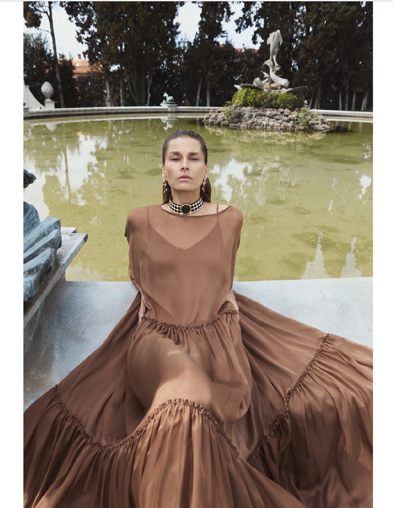 A woman in a brown dress poses beside a pond with a sculpture; she wears statement earrings and an embellished choker. The setting is outdoors.