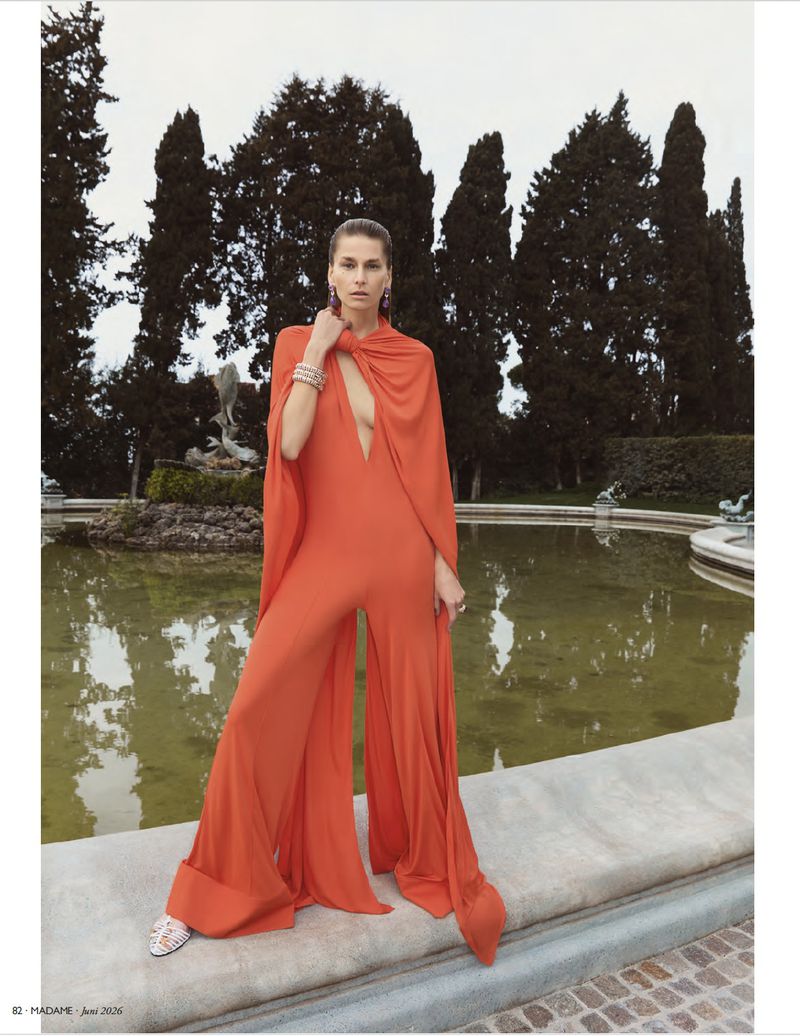 Model Hana Soukupova in an orange jumpsuit posing outdoors by a fountain and tall trees for Madame Germany.