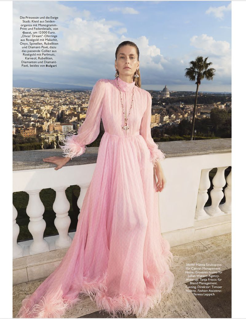 Model Hana Soukupova in a long pink dress standing on a terrace overlooking a city skyline under a partly cloudy sky.