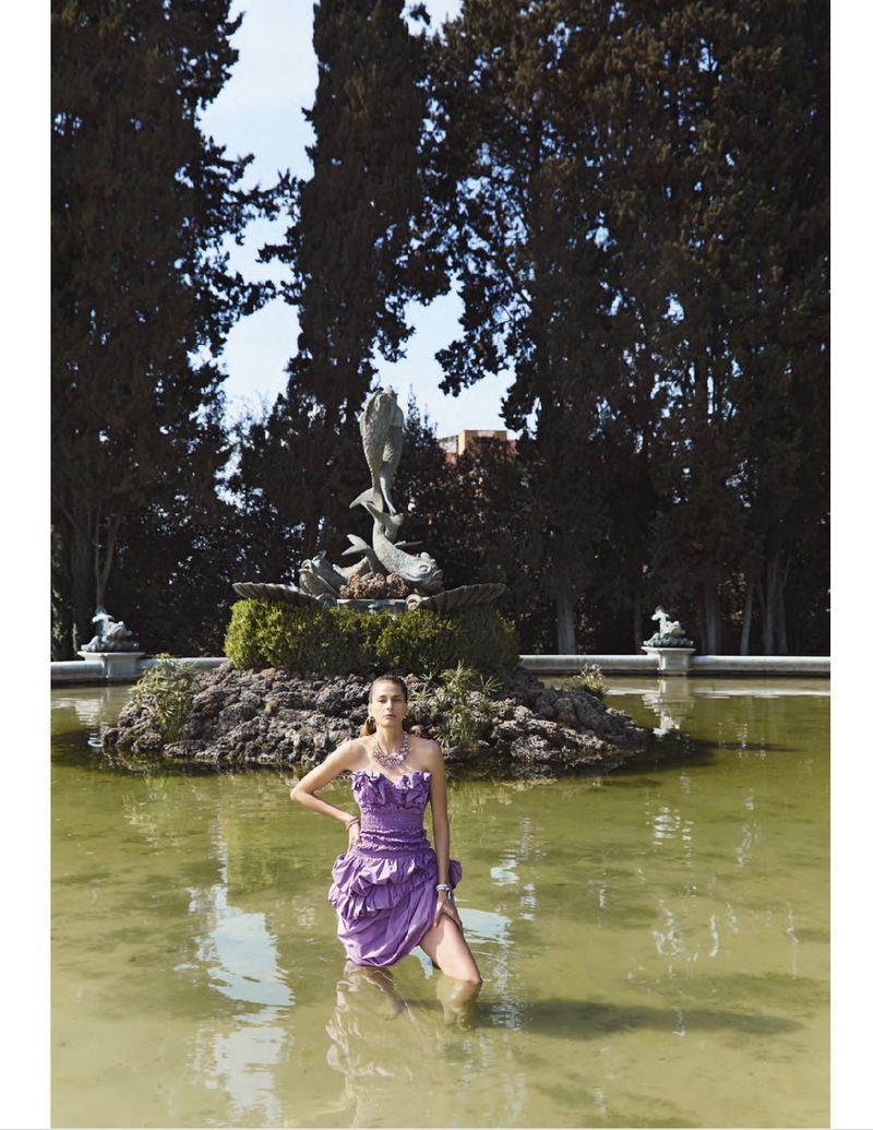 Model wearing a ruffled purple dress standing in a shallow outdoor fountain with a stone fountain sculpture in the background.