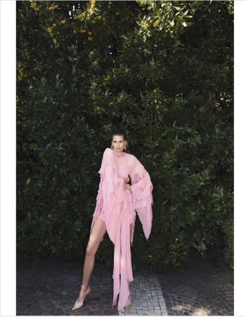 Model wearing a flowing pink ruffled dress standing on stone paving against a dense green foliage background.