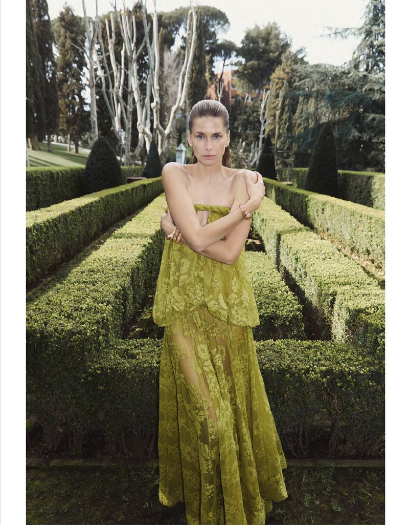 Model wearing a sheer yellow dress standing in a formal manicured garden with tall manicured hedges.