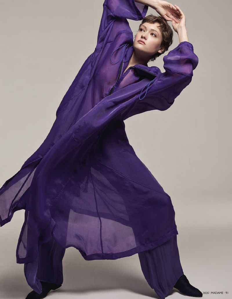 Model in a long sheer purple coat with hands raised in a dynamic pose against a neutral studio backdrop