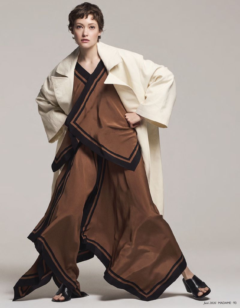 Fashion editorial shot of a young adult woman posing in a brown satin V-neck top and wide-leg pants with black trim, and a cream coat.