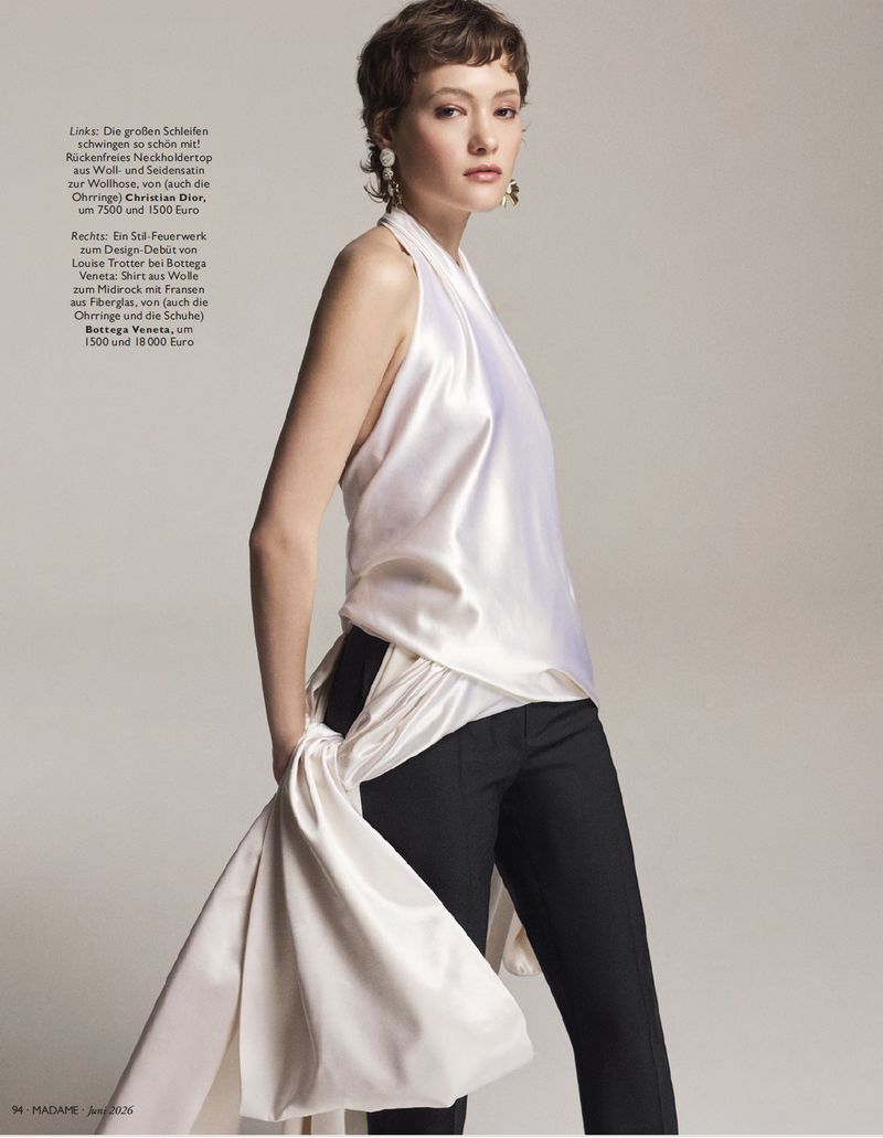 Model with short brown hair wearing a white silk halter top and dark trousers against a plain neutral background.