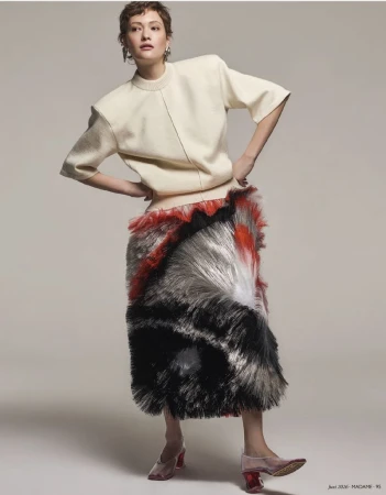 Model posing with hands on hips wearing a short-sleeved cream top and a multi-colored fringed skirt against a plain grey background
